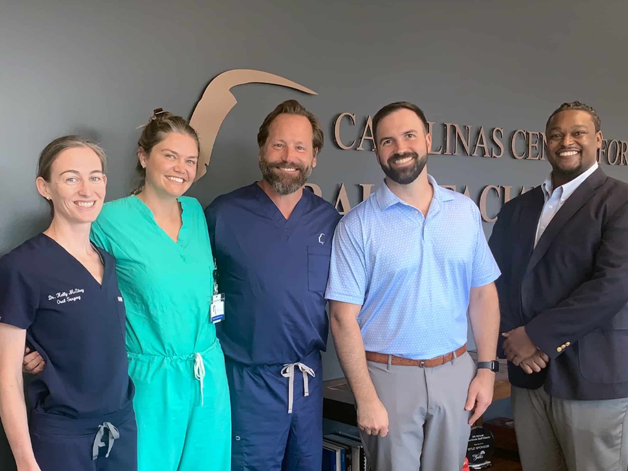 Five people smiling indoors, standing before a wall with "Carolinas Center for Oral & Facial" signage, wearing professional attire and scrubs.