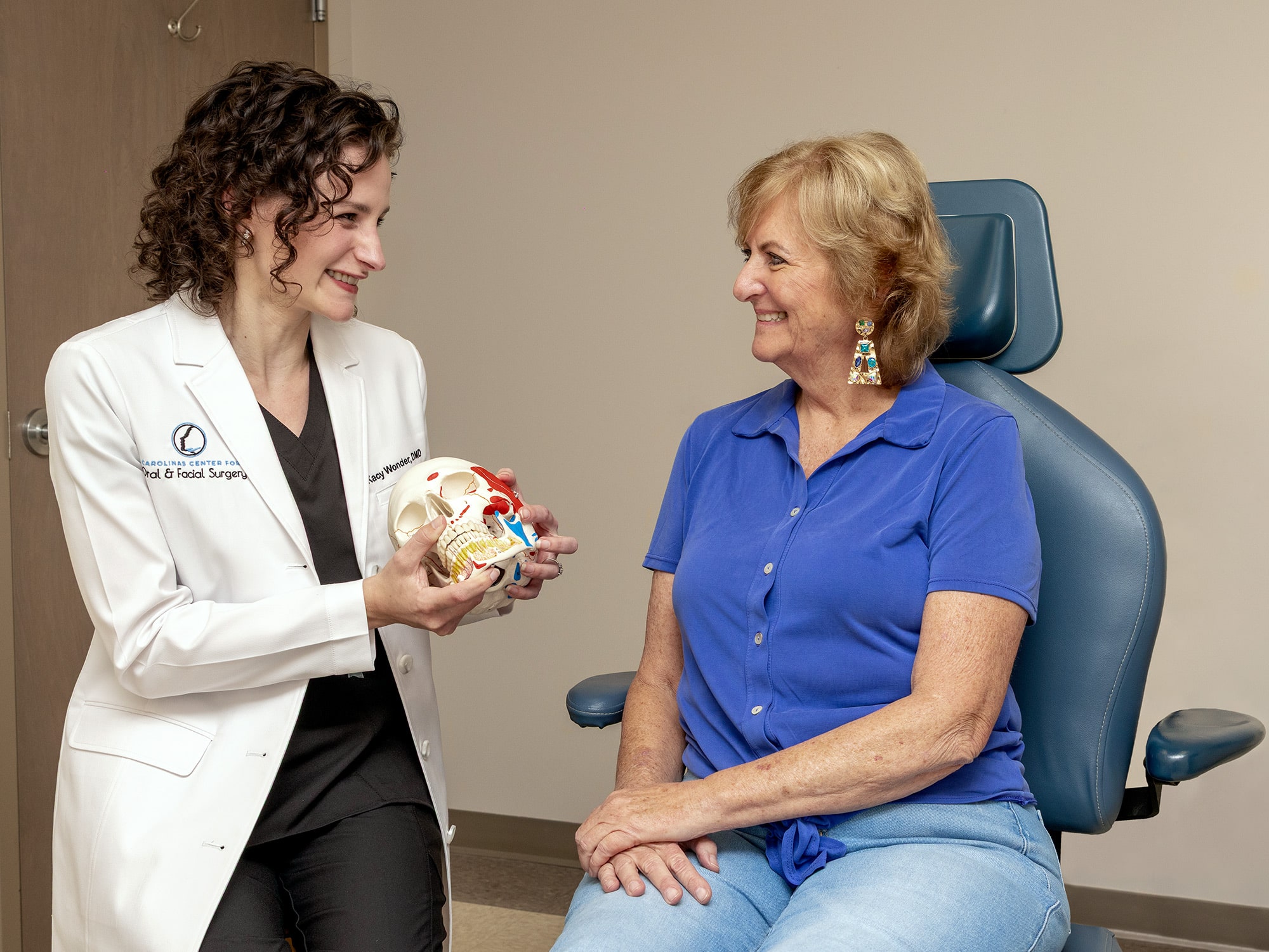 Two people are in a medical consultation. One holds a skull model, while the other sits attentively in a chair, smiling.
